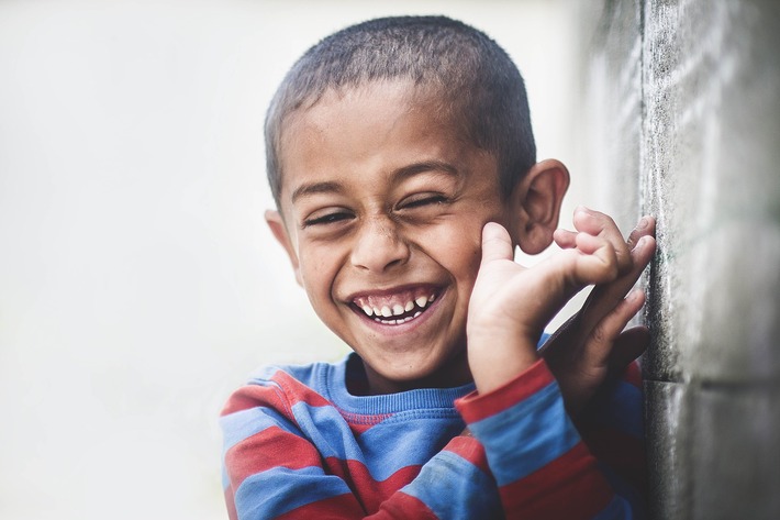 A young, smiling boy stands against a wall looking thankful and happy.