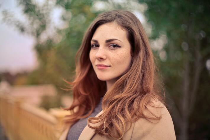 A young brunette woman stands on a dirt road looking at the camera with a small smile.